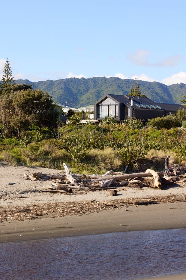 A modern dark-clad single-storey house with a steep gabled roof and large glazed gable end, set behind coastal vegetation on a sandy beach. Driftwood lies along the shoreline in the foreground, with calm water and a forested mountain range visible beyond the property.