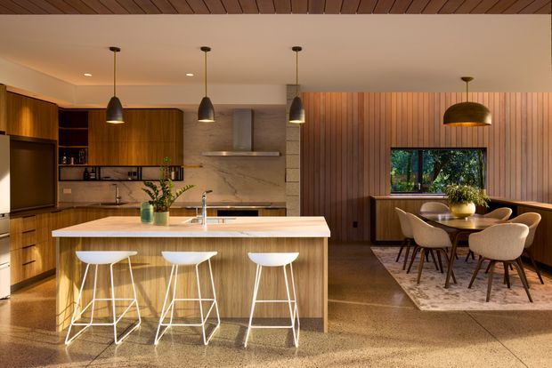 A warm, open-plan kitchen and dining area with extensive timber cabinetry and wall panelling. The kitchen features a large light stone island with three white bar stools, integrated appliances, open shelving, pendant lighting, and a marble-look splashback. Beyond the island is a dining zone with a timber table, upholstered dining chairs, a rug, and a large decorative pendant. The space has polished concrete flooring and a calm, refined mid-century-influenced aesthetic.