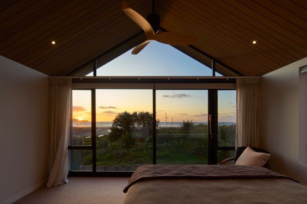 A warm, moody bedroom with a prominent vaulted timber-lined ceiling and exposed gable form. Full-height glazed sliding doors open the room to a sunset coastal or waterfront outlook, with curtains framing the view. A large bed occupies the foreground, and a ceiling fan hangs centrally above it.