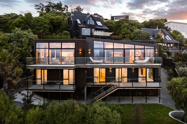 A multi-level contemporary residential house set into a lush, wooded sloping site. The dark-clad building features extensive floor-to-ceiling glazing, multiple balconies and terraces, a prominent external stair, and a mix of flat-roofed and pitched-roof volumes. Warm interior lighting contrasts with the charcoal exterior at dusk.