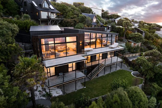 A dusk aerial view of a multi-level contemporary house on a densely planted, sloping suburban site. The home features dark vertical cladding, extensive black-framed glazing, multiple cantilevered decks and terraces, external stairs, and warm interior lighting. A smaller dark-clad house and neighbouring homes are visible among mature vegetation in the background.
