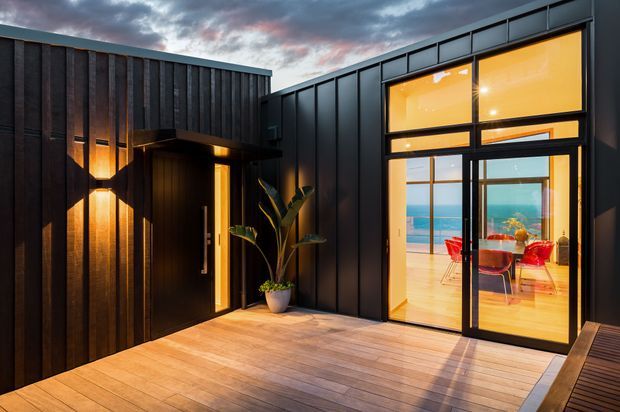 Exterior architectural view of a contemporary dark-clad coastal home at dusk. The composition focuses on a timber deck and recessed entry courtyard, with dramatic black vertical cladding, large black-framed glazed doors and clerestory windows revealing a bright dining or living area beyond. The ocean is visible through the glazing, while warm interior lighting contrasts with the dark exterior envelope and cloudy evening sky.