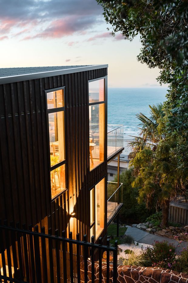 A narrow, multi-level contemporary coastal house viewed from an elevated garden path. The dark vertically clad facade has a low-pitched roof, tall warm-lit windows, glass-balustraded balconies, and black metal railings. Dense planting and stone retaining walls frame the building, with a clear ocean horizon beyond.