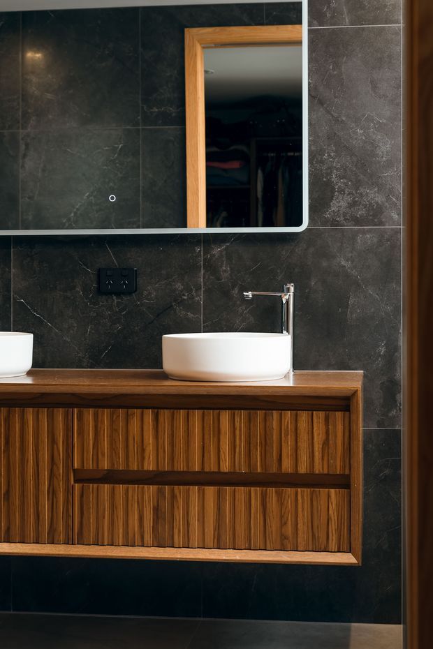 A contemporary bathroom vanity featuring a white vessel basin, chrome wall-mounted tap, dark marble-look tiled walls, and a richly grained timber vanity with vertically fluted cabinetry. A rounded-corner mirrored cabinet with a timber frame is mounted above the vanity.