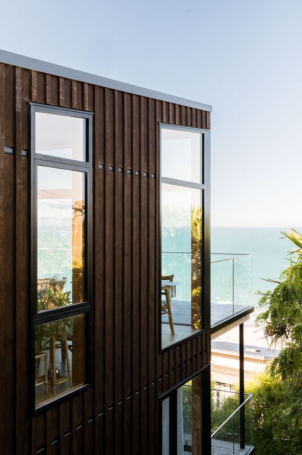 A contemporary multi-storey building facade clad in dark vertical timber boards, with tall black-framed windows reflecting and revealing interior spaces. Glazed balconies with slim dark railings project from the building, overlooking a bright coastal body of water. Lush planting surrounds the lower levels, creating a strong indoor-outdoor and elevated coastal setting.