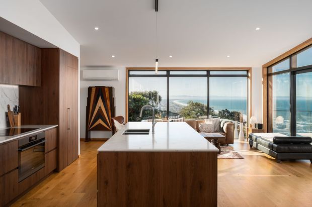 A bright open-plan kitchen and living area with a large walnut island, integrated timber cabinetry, white stone benchtops, and warm timber flooring. Floor-to-ceiling black-framed glazing opens to expansive ocean and coastal views, while a lounge and chaise occupy the living zone. The space has a clean contemporary composition with strong indoor-outdoor connection.