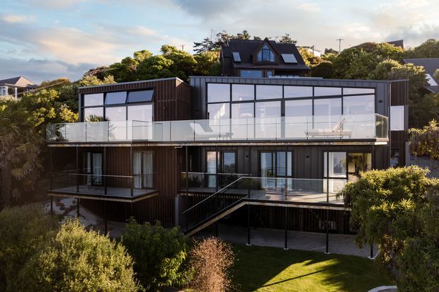 Exterior view of a contemporary multi-level residential building set among dense suburban landscaping. The dark-clad façade features extensive floor-to-ceiling glazing, projecting balconies with clear glass balustrades, roof-level windows, and an external stair leading to the elevated lower level.