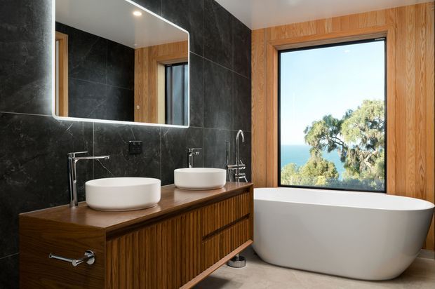 Contemporary bathroom featuring a double timber vanity with two white vessel basins, black marble-look wall tiles, chrome fixtures, a freestanding white bathtub, and warm timber window surrounds. A large window provides views toward trees and a blue body of water or sky.
