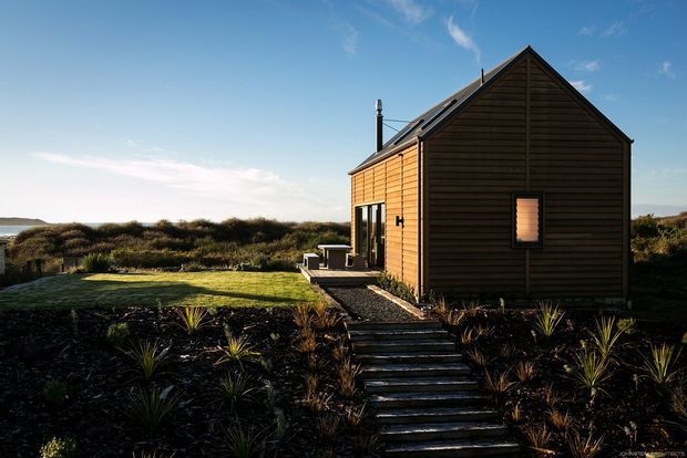 A small detached timber-clad dwelling set in a coastal-looking landscape, viewed from the exterior. The compact structure has a steep simple gable roof, dark horizontal weatherboard cladding on the end wall, warm timber side cladding, a narrow glazed door, small windows, and a stepped path leading through native-style planting to the entrance. Clear blue sky and low scrub surround the building.