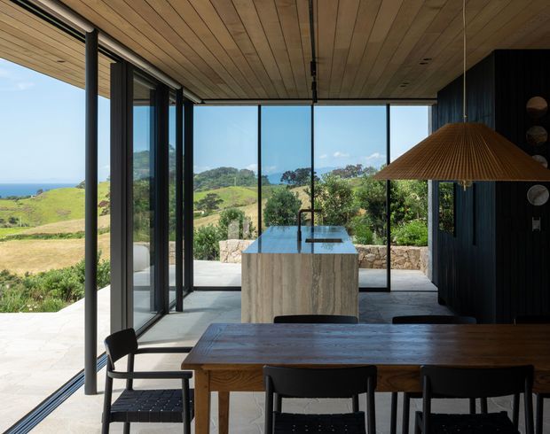A contemporary open-plan dining and kitchen interior with extensive floor-to-ceiling glazing overlooking a green coastal landscape. The room features a long dark timber dining table with black chairs, a central stone island, dark vertical timber wall cladding, a warm timber-lined ceiling, and a large woven pendant light. Black-framed glass doors open toward an outdoor terrace, creating a strong indoor-outdoor connection.