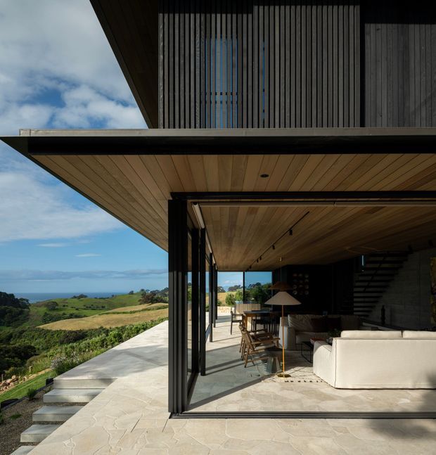 A contemporary multi-level home with broad cantilevered roof planes, dark vertical timber screening, expansive retractable glass openings, and warm timber soffits. The covered outdoor living area contains lounge and dining furniture and opens onto a large pale stone terrace with steps. Rolling green coastal landscape and ocean are visible beyond the house.