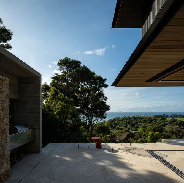 A contemporary open-air terrace overlooking dense coastal vegetation and the ocean. The space features broad pale stone paving, a built-in outdoor fireplace or barbecue clad in rough natural stone and dark concrete, sculptural outdoor chairs, and dramatic cantilevered timber-lined roof eaves. The view is framed by mature trees, with a calm blue sky and distant shoreline beyond.