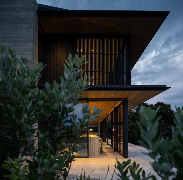 Twilight exterior view of a contemporary multi-level residence, framed by dense planting. The building features strong horizontal roof overhangs, dark vertical screening, extensive glazing, warm timber soffits, and a sheltered ground-floor outdoor entertaining or circulation area with a visible bar or joinery element. The composition emphasises the dramatic roof forms, indoor-outdoor connection, and warm architectural lighting.