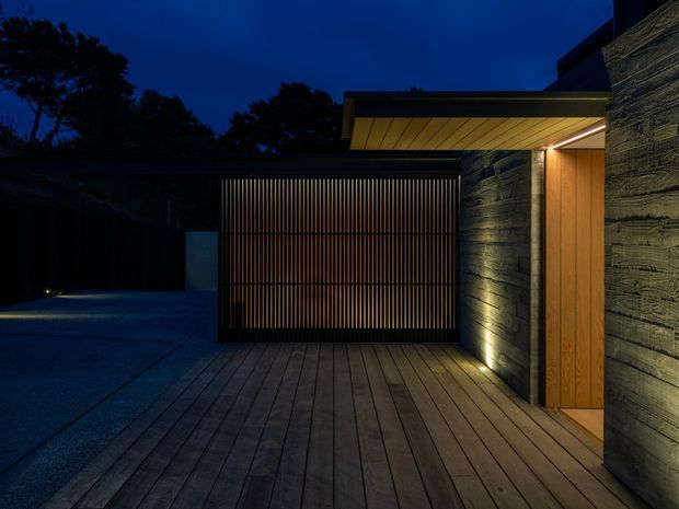 A dramatic dusk exterior view of a contemporary residential entry and garage area. A warm timber-clad doorway sits beneath a projecting flat roof, beside textured dark masonry walls and a tall vertical timber slat garage screen. The entry and garage open onto a broad timber deck, with integrated low-level lighting creating a warm contrast against the deep blue evening sky and shadowed landscaping.