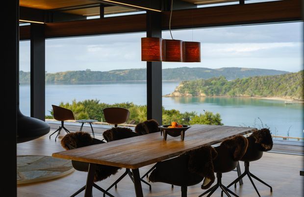 A contemporary dining area with a long solid timber dining table and dark upholstered dining chairs, positioned in front of expansive floor-to-ceiling glazing overlooking a calm waterfront landscape. The room features dark structural framing, a suspended linear light, and a cluster of warm timber pendant shades, creating a refined indoor-outdoor connection.