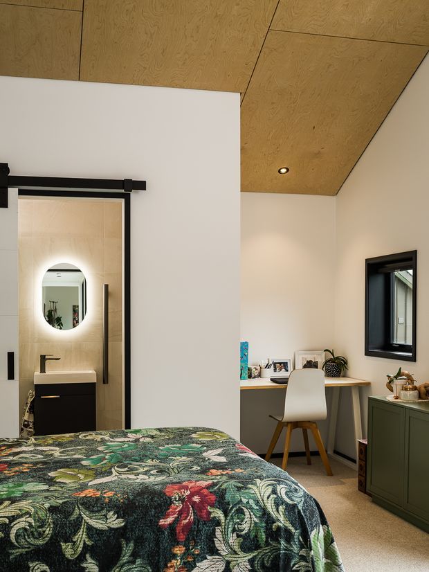 A compact, warmly styled bedroom with a richly patterned green and floral bedspread in the foreground. The room features a pale neutral wall, a timber-panelled ceiling with recessed downlights, black-framed windows and a black barn-style sliding door. Through the open doorway is a small bathroom with a wall-mounted black vanity, beige tiled walls, a backlit oval mirror and black tapware. A simple desk with a white chair sits beneath the window, alongside green built-in cabinetry and decorative objects.