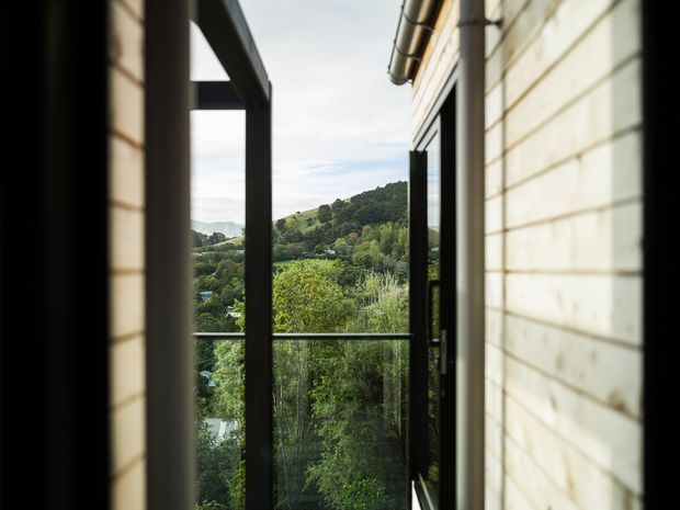 A narrow exterior passage or balcony beside a light-colored weatherboard-clad building, with dark-framed glazing and a glass balustrade opening toward a lush, hilly landscape. The image emphasizes the building edge, outdoor circulation space, and green suburban or rural outlook in soft natural light.