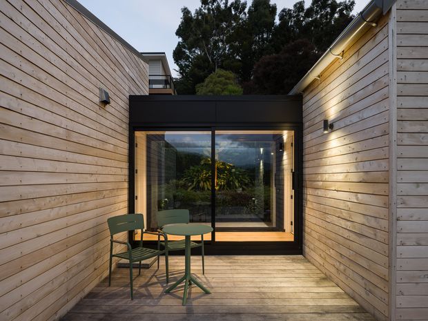 A compact enclosed timber courtyard or deck between two horizontal timber-clad walls. The space features timber decking, a black-framed glazed doorway or window, warm integrated wall lighting, and a small green outdoor table and chairs. The glazing reflects and reveals greenery beyond, while the upper roofline and surrounding trees suggest a private residential outdoor retreat.