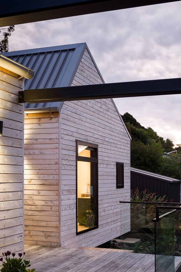 Exterior architectural photograph of a contemporary timber-clad building with a steep gabled metal roof. Pale horizontal weatherboard cladding contrasts with dark window frames and structural beams. A timber deck with glass balustrading occupies the foreground, with landscaped vegetation and a dark outbuilding visible behind. Warm interior light glows through the tall narrow windows under an overcast sky.