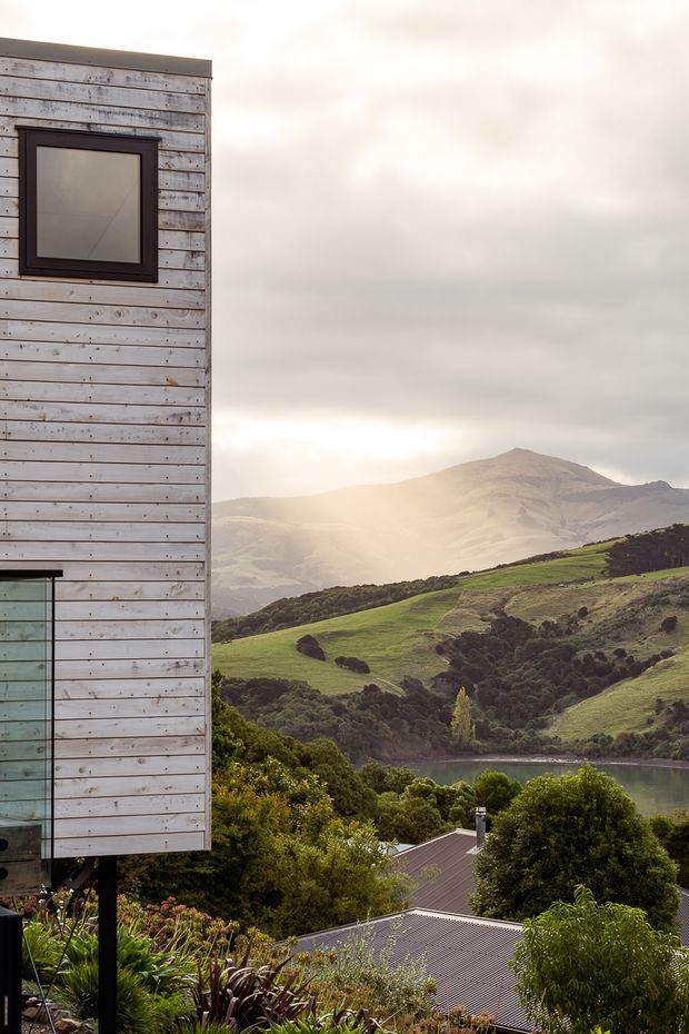 A vertical exterior architectural view showing the side of a modern elevated building clad in weathered horizontal timber boards, with dark-framed square glazing and a large window or glazed opening at the lower left. The building overlooks a lush, hilly landscape with a lake or river inlet and a mountain beyond under soft overcast light. Roofs and dense planting are visible below.