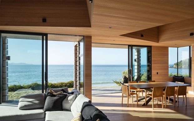 A bright, open-plan coastal living and dining space with expansive floor-to-ceiling glazing overlooking the ocean. The room features extensive warm timber wall and ceiling lining, a sculptural timber ceiling recess, dark-framed glass doors and windows, a large light-coloured sectional sofa, and a timber dining table with matching chairs. An outdoor terrace and planted coastal landscape are visible beyond the open glazing.