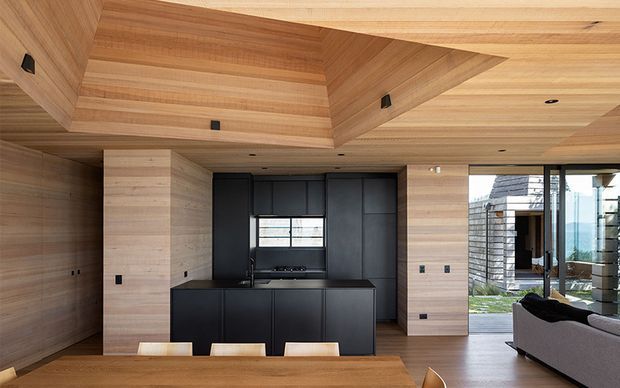 A refined open-plan kitchen and living area featuring extensive light timber wall, ceiling, and built-in cabinetry finishes. The kitchen has a matte black island and dark cabinetry, integrated appliances, and a compact horizontal window. A sculptural recessed timber ceiling detail adds architectural interest, while full-height glazing opens toward an outdoor area and brings in natural light. The foreground includes a timber dining table, and part of a grey sofa is visible to the right.