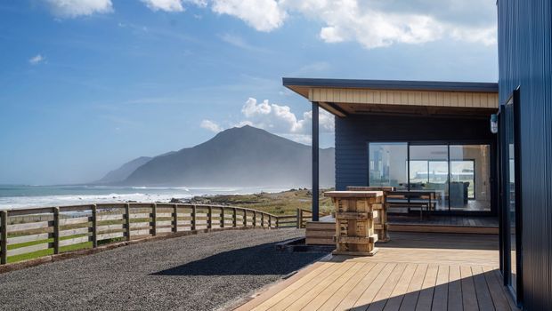 A contemporary coastal building viewed from a timber deck, with dark vertical cladding, a simple flat-roofed veranda, large glazed sliding doors, and an outdoor timber bar or dining bench. The property overlooks a sandy beach and ocean with a prominent mountain in the background. Gravel landscaping, timber fencing, and decking create a relaxed outdoor setting.
