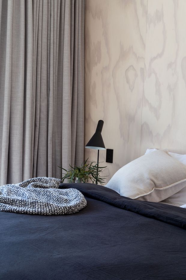 A close-up of a softly styled contemporary bedroom. The bed is dressed in dark charcoal bedding with a textured black-and-white throw and large pale cushions. A raw, pale timber feature wall sits behind the bed, alongside full-height taupe-grey curtains. A black wall-mounted reading lamp and small leafy plant add contrast and texture.