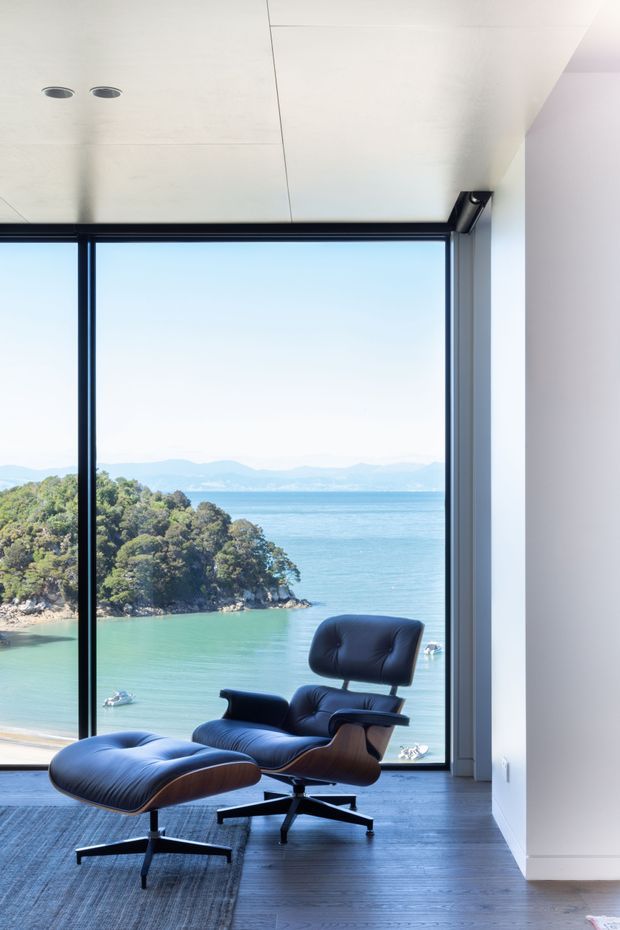 A bright, minimalist interior with a black-framed floor-to-ceiling window overlooking a blue coastal inlet and tree-covered shoreline. A dark leather lounge chair and matching ottoman sit on a dark timber floor beside a textured rug. The room has a smooth white ceiling with recessed downlights and plain white walls.