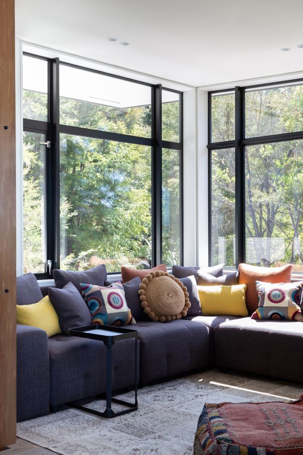 A bright, contemporary living room with a large dark upholstered sectional sofa arranged beside expansive black-framed corner windows. The room has white walls and ceiling, abundant natural light, colourful patterned cushions, a round textured accent cushion, a small black side table, and layered patterned rugs. Lush greenery is visible outside through the glazing.