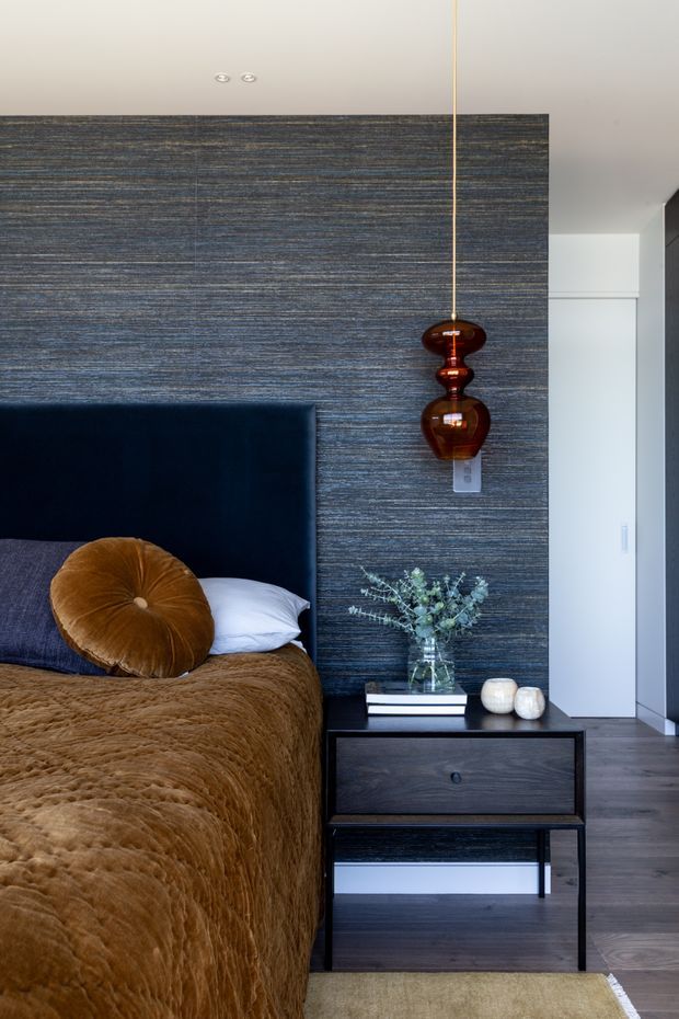 A refined contemporary bedroom featuring a dark navy upholstered bedhead, richly textured blue-grey wall covering, and a brown velvet bedspread with matching round cushion. A dark timber bedside table supports books, a glass vase of foliage, and ceramic vessels. A sculptural amber glass pendant hangs beside the bed, with dark timber flooring and a neutral rug completing the layered, moody scheme.