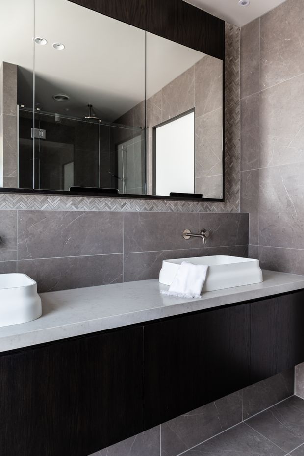 Contemporary bathroom vanity with a long pale stone benchtop, dark timber cabinetry, a white vessel basin, wall-mounted brushed metal tapware, and large mirrored cabinetry above. Grey stone-look wall and floor tiles are accented by a narrow herringbone tile detail beneath the mirror. A frameless glass shower enclosure is visible in the reflection. The space has a refined, monochromatic and slightly moody finish.