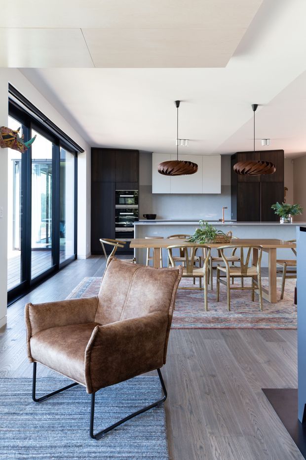 A bright, open-plan contemporary interior combining a living area, dining area and kitchen. The foreground features a brown leather lounge chair on a blue-grey rug, while the dining zone has a timber table and sculptural timber chairs beneath two dark wood pendant lights. The rear kitchen includes a large island with pale cabinetry, dark timber appliance and storage towers, a grey backsplash, and a copper-toned tap. Full-height black-framed sliding glass doors open to an outdoor deck, and the room has a light timber ceiling feature and dark timber flooring.