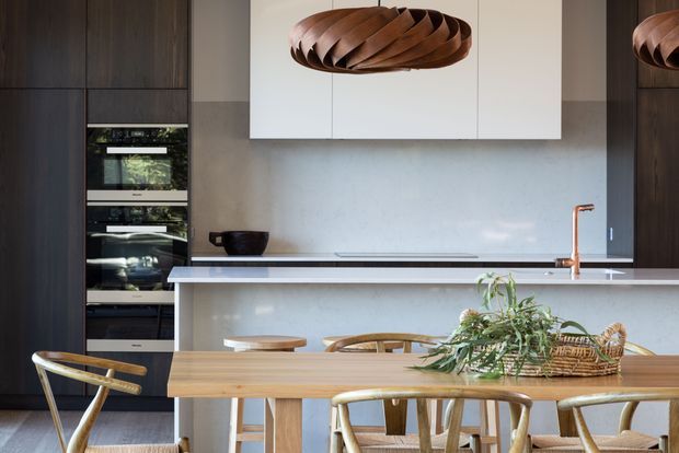Contemporary kitchen and dining area with dark timber cabinetry, integrated appliances, a pale stone island benchtop, and a timber dining table with woven timber chairs. Sculptural wood pendant lights and natural greenery add warmth to the refined, neutral interior.