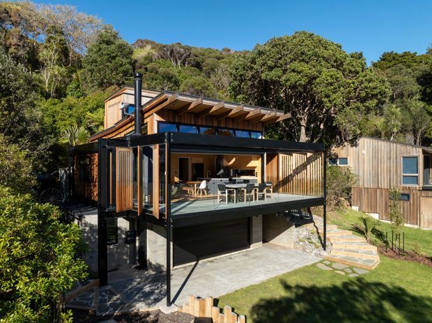 Exterior view of a compact multi-level timber-clad house set among dense vegetation. The home features a projecting upper deck with dark metal framing and glass balustrades, an open-plan living and dining area behind large glazing, a covered outdoor terrace, a skillion-style roof with exposed timber eaves, and a lower-level garage or storage area. A separate timber-clad structure and landscaped paths are visible to the side.