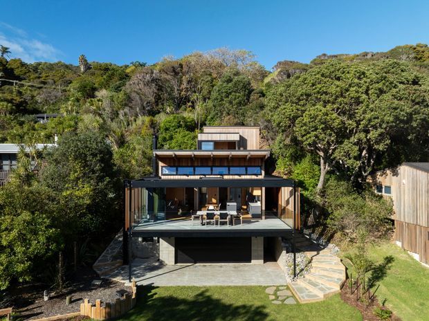 Exterior view of a contemporary multi-level house set into a densely vegetated, sloping site. The home has dark framed floor-to-ceiling glazing, projecting balconies, timber-clad upper volumes, a flat roofline, and a lower-level garage or covered undercroft. Stone steps and concrete paving connect the house to a landscaped lawn.