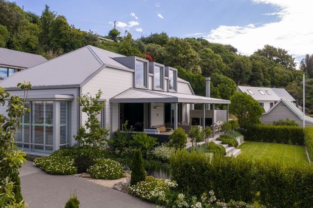 Exterior view of a light-coloured, multi-level suburban house set into a lush, landscaped site. The home has a steep gabled roof with grey metal roofing, horizontal light weatherboard cladding, projecting dormer windows, and a covered veranda or porch. A neighbouring gabled house is visible to the right. Formal hedges, lawns, planted garden beds, and a paved walkway frame the foreground.