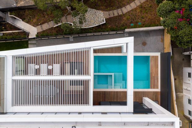 Aerial view of a contemporary white-roofed residence with a geometric rooftop terrace and vivid turquoise swimming pool. The roof incorporates timber decking, white parapet walls, and a large slatted pergola or screened outdoor area. A landscaped garden with curved stone paths surrounds the property.
