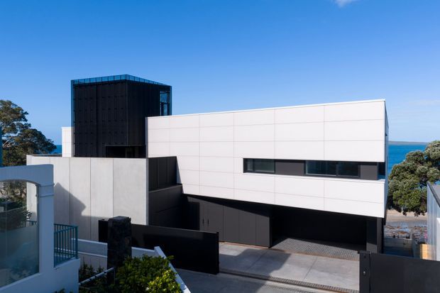 Exterior view of a sharply geometric multi-level contemporary residence, featuring a white horizontal upper volume, dark charcoal lower walls and garage opening, and a tall black vertical glazed or screened element. The property is viewed from an elevated street or neighbouring position, with the ocean visible beyond and mature coastal vegetation around it.