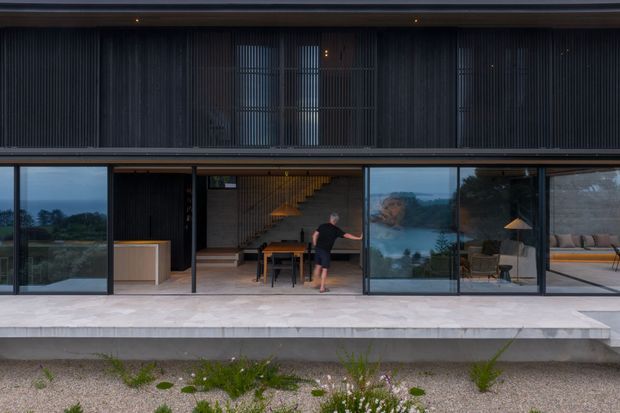 Exterior view of a contemporary two-storey residence with a dark, finely ribbed upper facade and extensive ground-floor glazing. Sliding glass doors open onto a broad pale stone terrace, revealing a dining area, sculptural staircase, and living space inside. The home appears elevated above a gravelled landscaped foreground, with a calm, refined indoor-outdoor connection.
