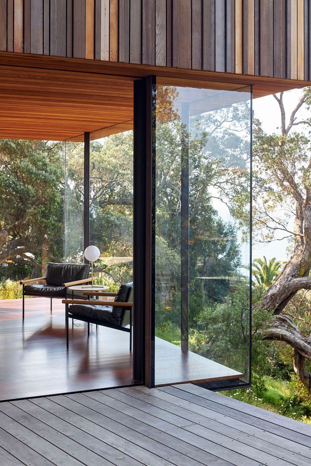 A modern indoor-outdoor living space with extensive floor-to-ceiling glazing opening onto a timber deck in a wooded setting. The interior features a warm timber-lined ceiling and wall, dark-framed glass panels, polished dark timber flooring, and black leather lounge chairs with slender timber arms. The architecture emphasises a strong connection to the surrounding landscape.
