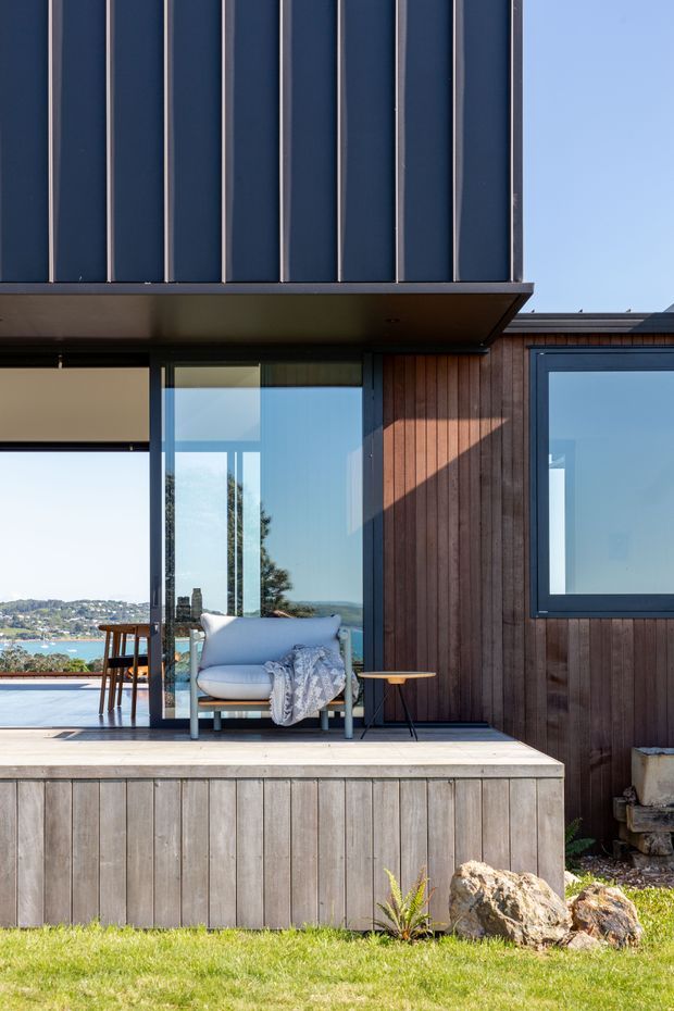 Exterior view of a contemporary timber-clad house with dark vertical metal cladding, large glazed openings, and a raised timber deck overlooking a visible coastal bay. A cushioned outdoor armchair and small side table sit on the deck, with landscaped rocks and lawn in the foreground.