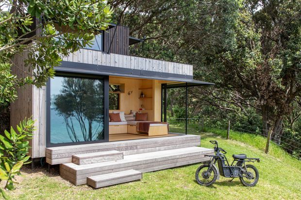 A compact contemporary timber-clad dwelling nestled among mature trees, viewed from the garden. The elevated structure has broad glazed openings, a covered outdoor living area, and wide stepped decking or terrace stairs leading to the lawn. Warm timber interiors, built-in seating, and a bicycle are visible through the open glazing.