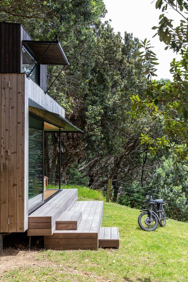 Exterior view of a compact contemporary timber-clad dwelling nestled among dense native vegetation. The building has vertical dark and natural timber cladding, expansive glazing, projecting angled awnings, and a stepped timber deck with integrated stairs. A wire balustrade follows the edge of the elevated deck, while a bicycle rests on the lawn beside the house.