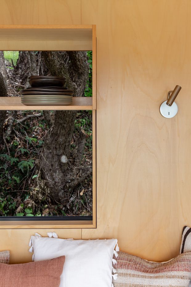 Close-up interior view of a warm, wood-lined bedroom featuring a built-in timber shelving niche with stacked bowls, a large window looking onto a tree, a simple wall-mounted reading light, and layered textured bedding in neutral and terracotta tones.