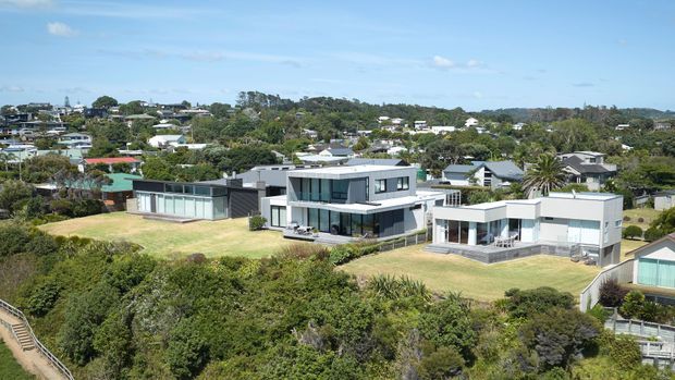 Aerial view of a large contemporary residential property on a landscaped coastal or elevated site, with multiple interconnected flat-roofed volumes, extensive glazing, dark and light exterior cladding, and broad lawns. A suburban neighbourhood and dense greenery are visible in the background.