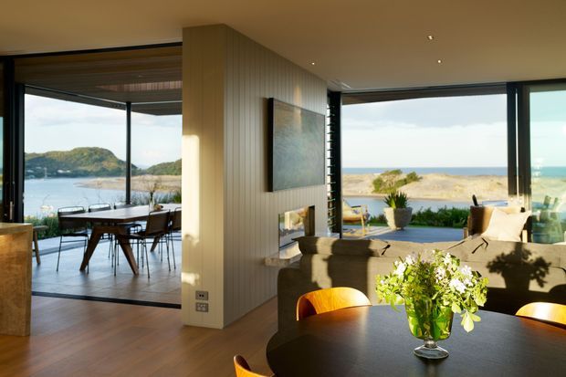 A refined open-plan coastal living and dining space with expansive floor-to-ceiling glazing overlooking a bay and headland. The room features warm timber flooring, a vertically panelled central partition with a fireplace and artwork, a dark rounded dining table with timber chairs, and a comfortable living area opening onto an outdoor terrace. The interior is flooded with natural light and strongly connected to the landscape.