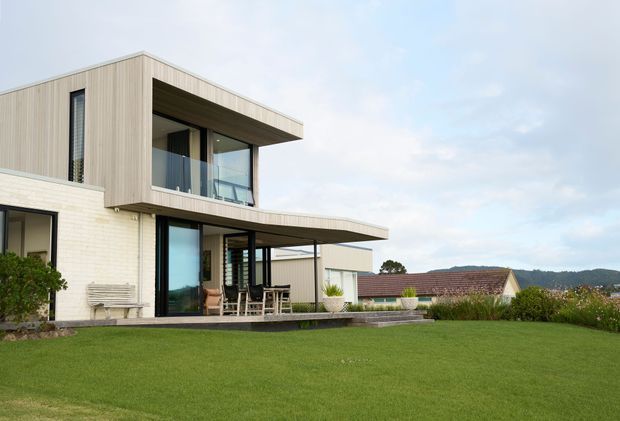 Exterior view of a modern two-storey home with a pale brick and vertical timber-clad facade, strong horizontal flat-roof overhangs, large glazed openings, and upper-level balconies. The house opens onto a broad manicured lawn with a paved terrace and outdoor dining area; neighbouring rooftops and wooded hills are visible in the background.
