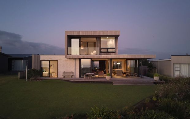 A contemporary two-storey residential house photographed at dusk. The home has a strongly rectilinear composition, extensive floor-to-ceiling glazing, warm timber-toned upper-level cladding and framing, pale masonry walls, and a broad flat roofline. A timber deck with outdoor seating runs along the ground floor, opening onto a lawn and planted garden.