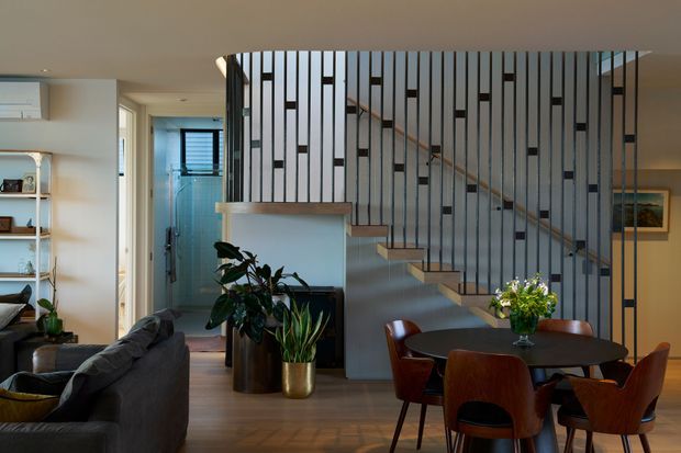 A contemporary open-plan living and dining area with a floating timber staircase enclosed by a distinctive black vertical metal balustrade and white panels. The room includes a dark sofa, round dining table with sculptural timber chairs, indoor plants, built-in shelving, timber flooring, and warm neutral finishes. A hallway and bathroom or utility area are visible beyond the living space.